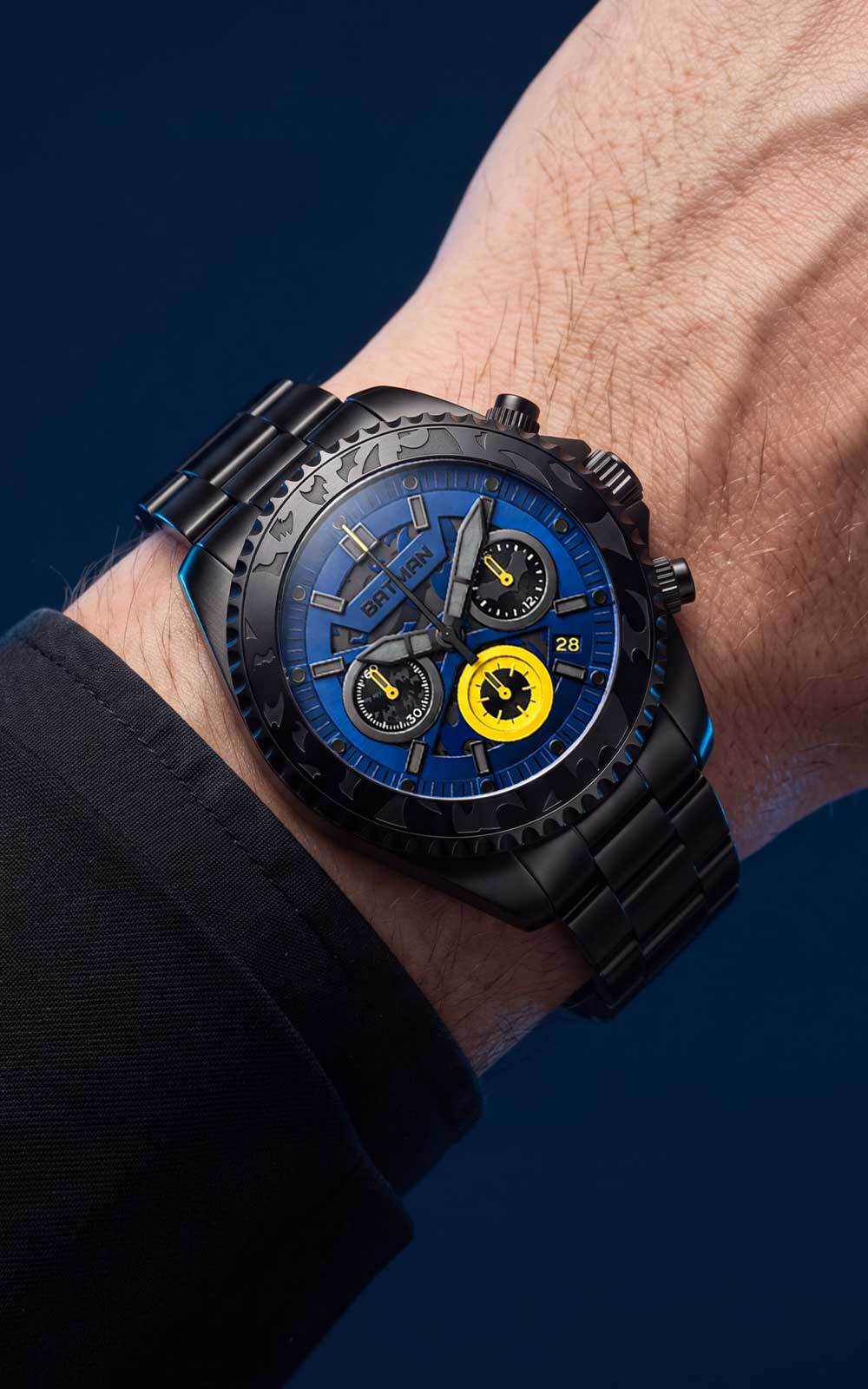 A close-up of a wrist wearing a black chronograph watch with a vibrant blue dial and a bright yellow subdial, set against a dark blue background.